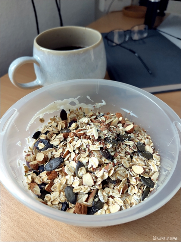 Schüssel mit Müsli und einer Teetasse, im Hintergrund liegt ein Notizbuch mit einer Lesebrille. Für die Aktion Zwölf von Zwölf im Juli