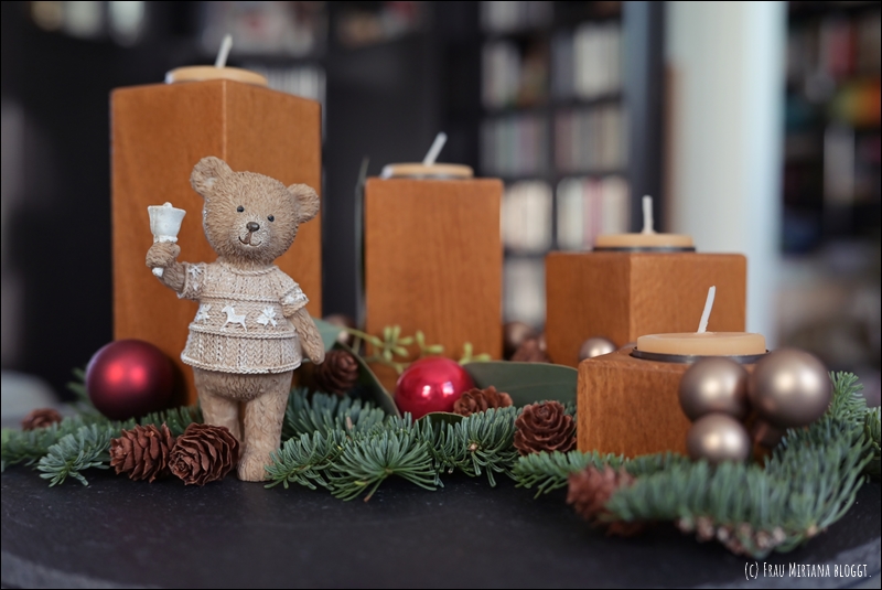 Kleine Teddybär-Figur mit Glocke in der Hand, vier Holzklötze in honigfarbenem Holz mit Teelichtern, dekoriert mit Tannengrün, roten und goldenen kleinen Kugeln und MIni-Tannenzapfen.